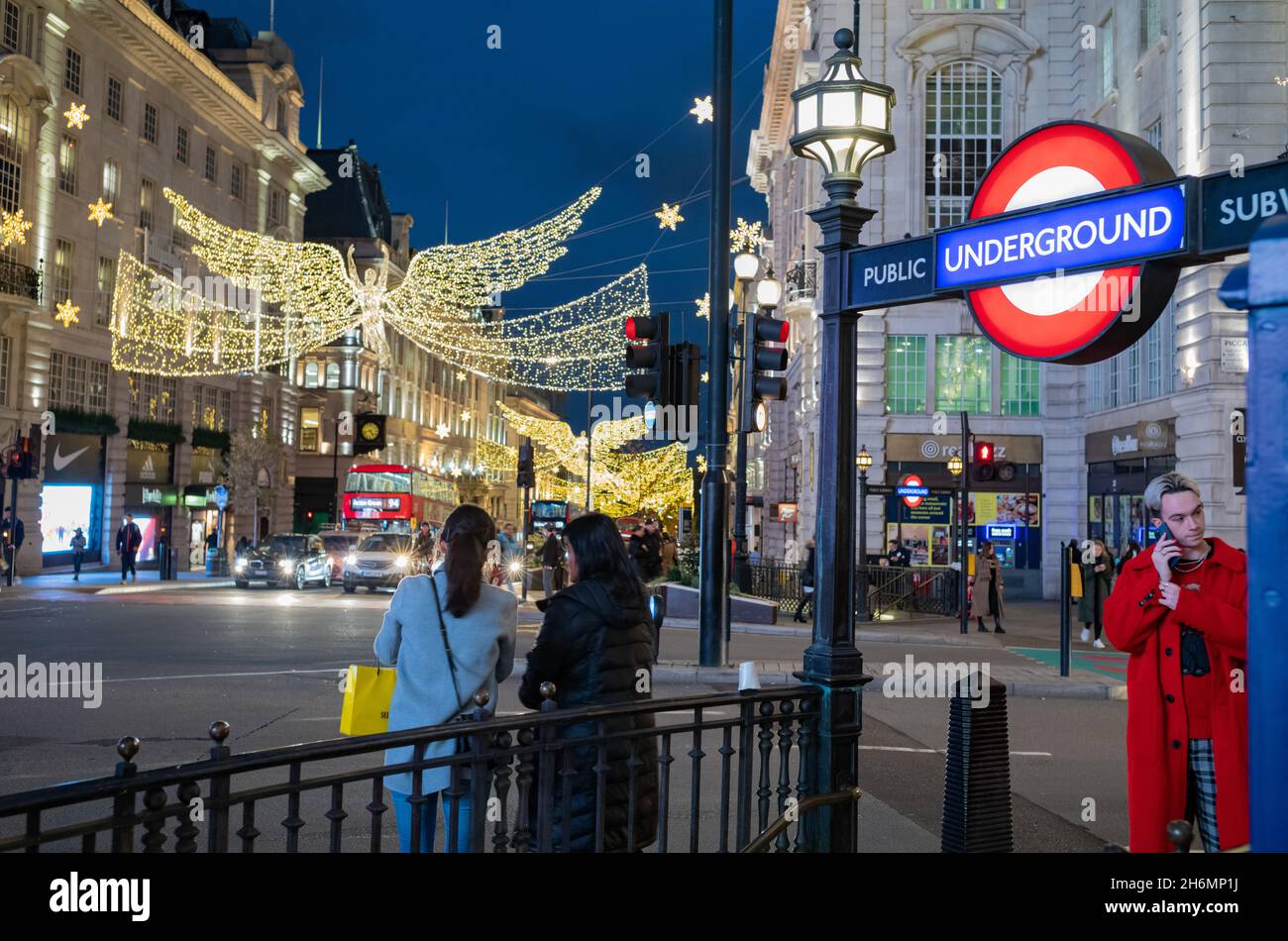 A yound man in a red coat speaks on the telephone at the entrance to Piccadilly Circus Tube Station as Christmas Lights illuminate the night streets Stock Photo