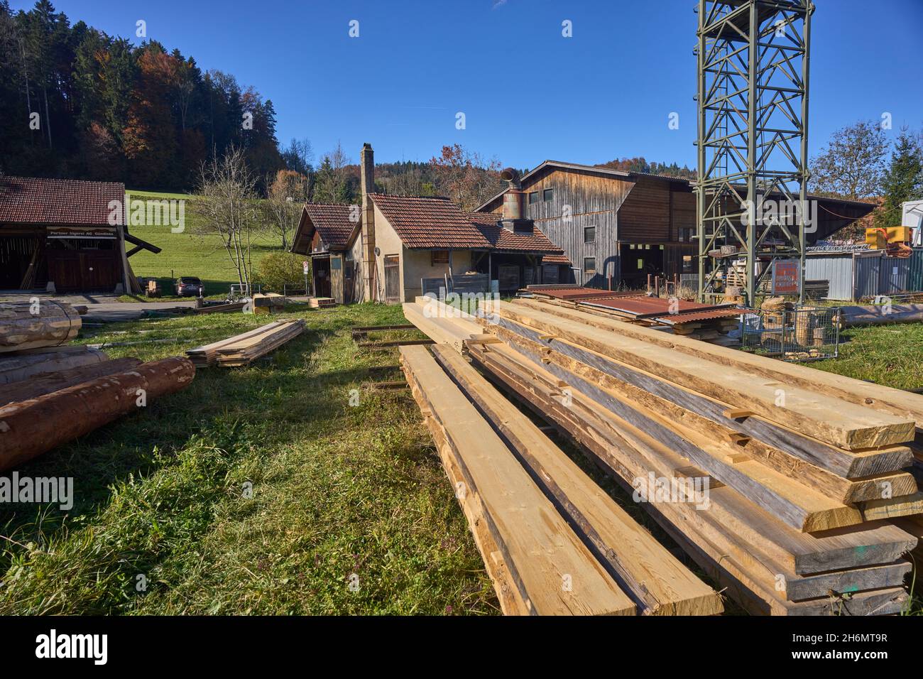 Outdoor Lumber Yard. In The Background Crane, Sawmill Building, Meadow, Forest And Blue Sky. Gündisau, Zurich Oberland, Switzerland Stock Photo