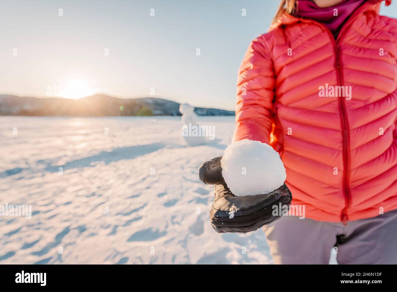 Winter outdoor active lifestyle concept. Girl holding snowball on snowy day with snowman in background on frozen lake at beautiful winter sunset Stock Photo