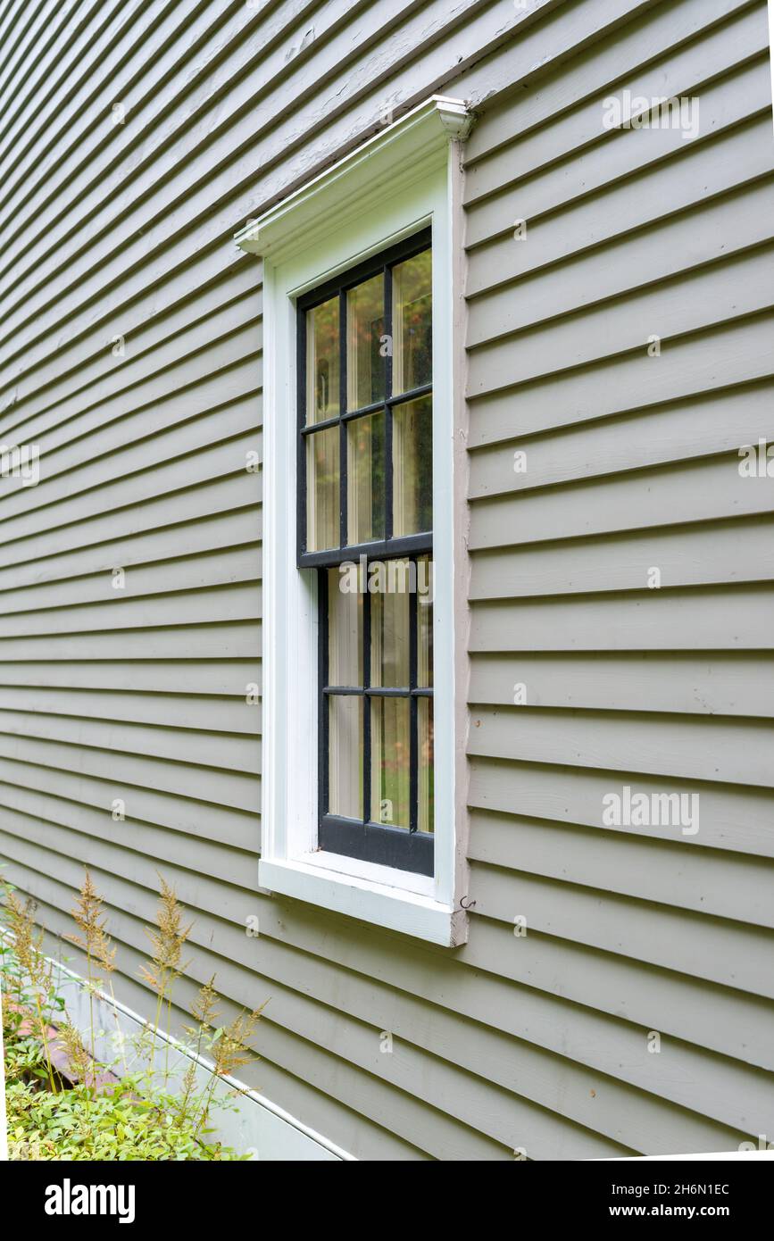 Old tan colored house with white trim and black wood window frame spacers. The glass in the window is wavy and reflects the sun. Stock Photo