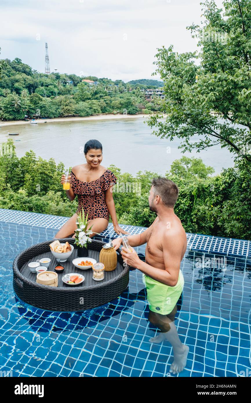 couple man and woman having Floating Breakfast tray in luxury pool hotel, young female with hat enjoy in a tropical resort. Relaxing, Exotic summer travel, holiday, vacation, and weekend. Stock Photo