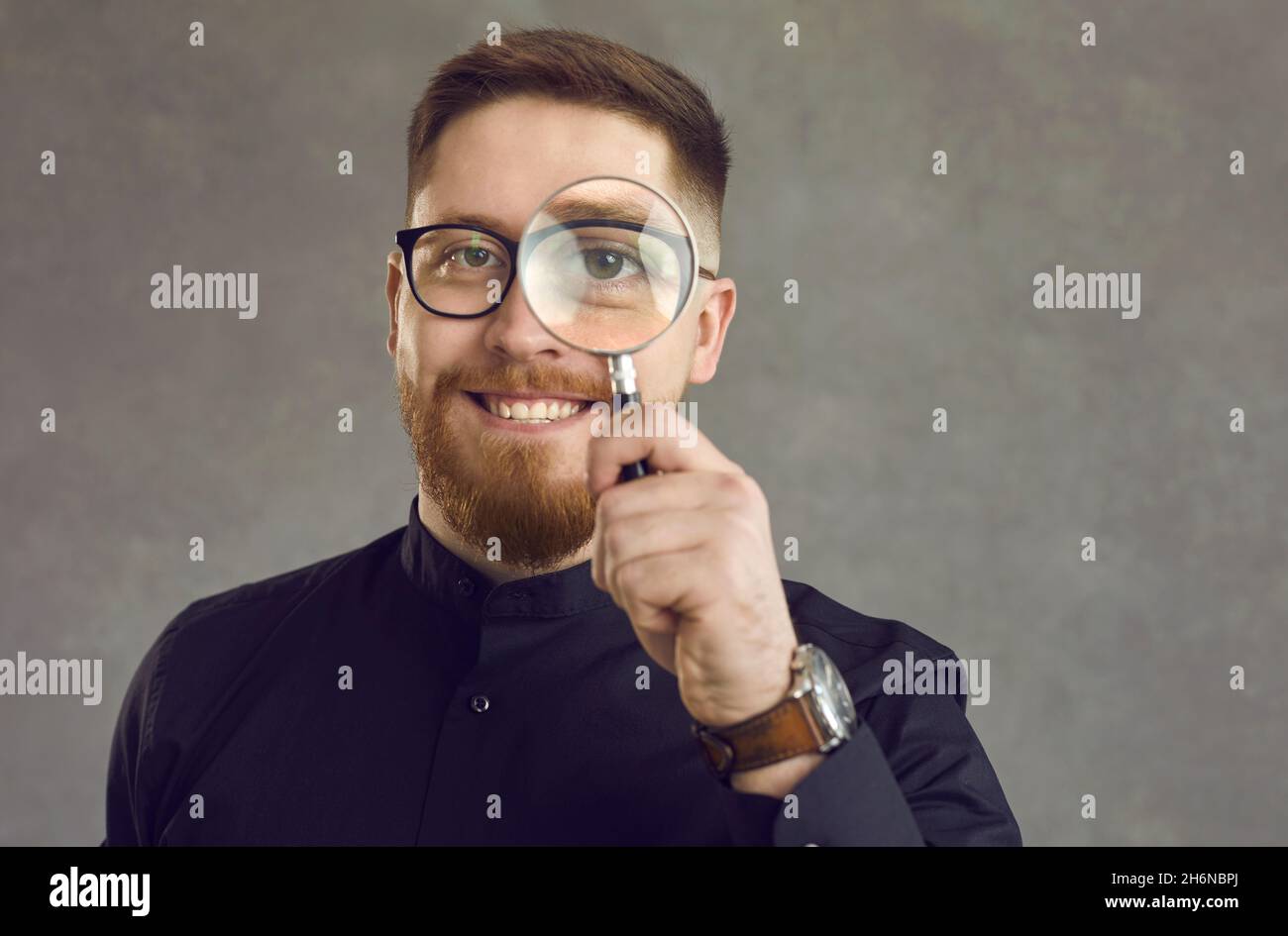 Positive young detective man looking through a magnifying glass while standing on gray background. Stock Photo