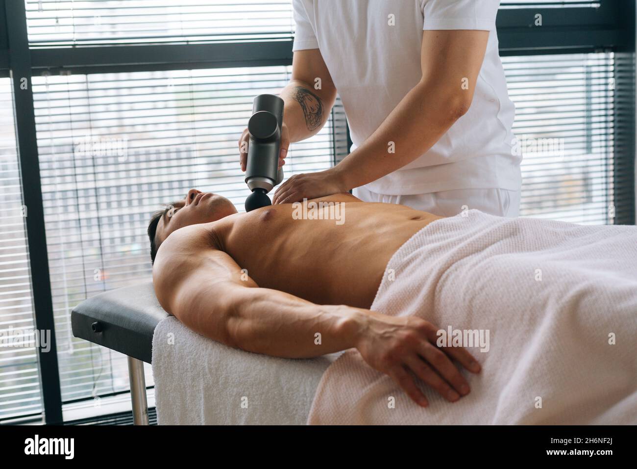 Medium shot of unrecognizable professional male masseur receiving chest muscles using massage gun percussion tool of muscular athlete man Stock Photo