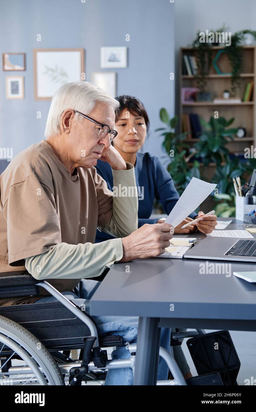 Senior man with disability reading a document at the table with laptop, he studying together with his teacher at home Stock Photo