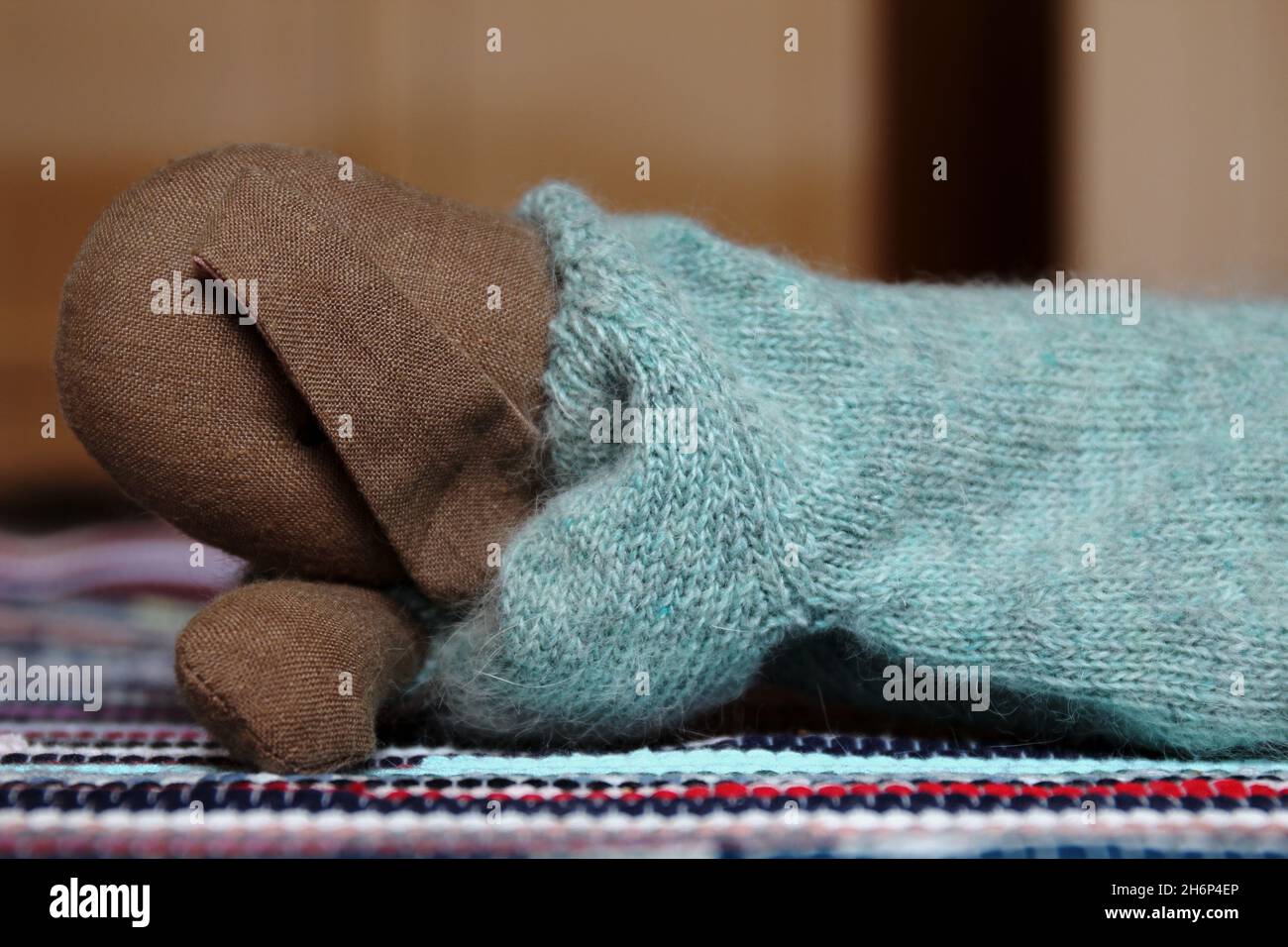 Kuscheltier süßer Hase mit langen Ohren und hellblauem Wollpulli, liegend auf einem bunten Teppich. Daneben eine rosa farbene Maske (Mundschutz). Stock Photo