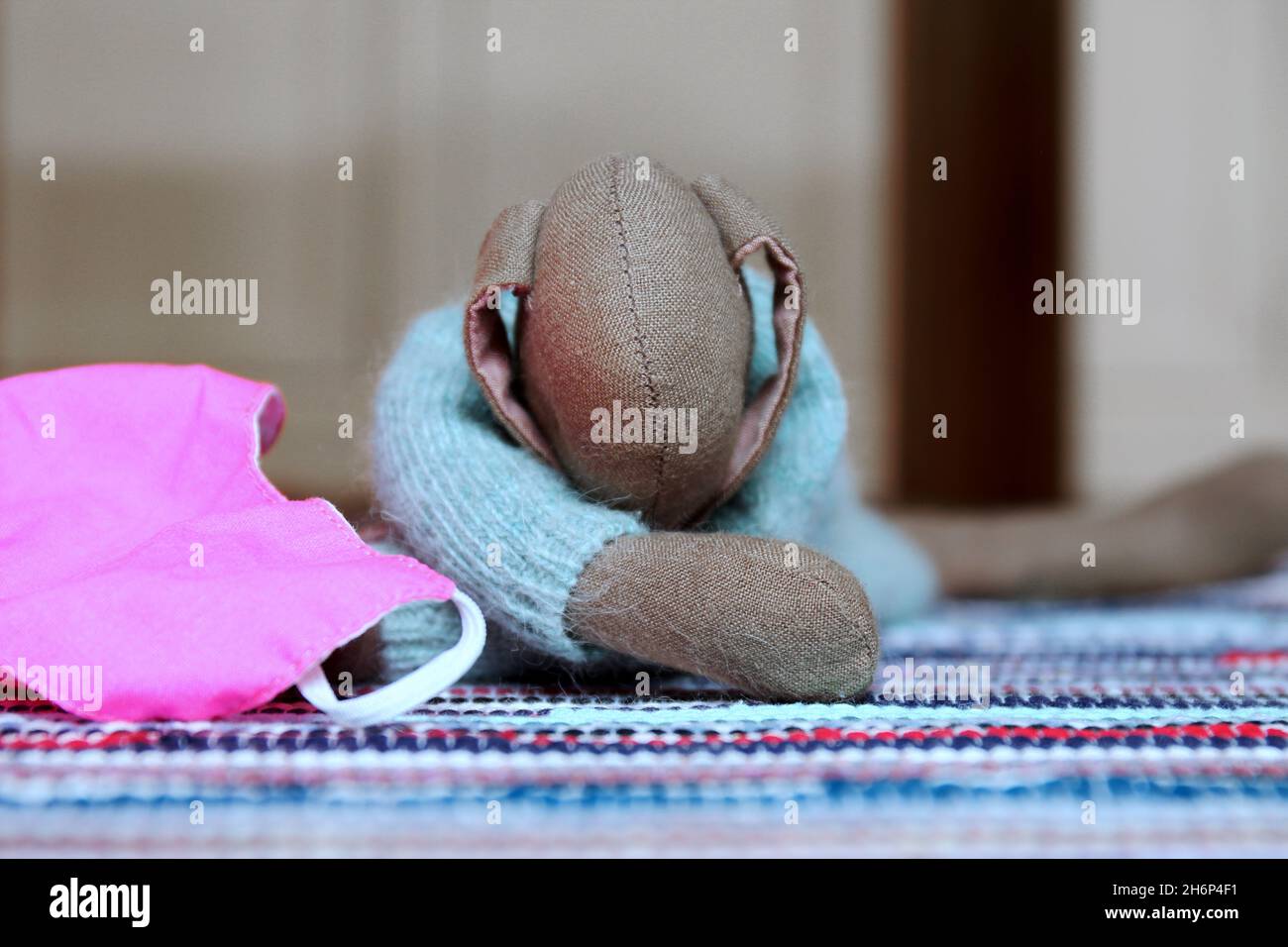 Kuscheltier süßer Hase mit langen Ohren und hellblauem Wollpulli, liegend auf einem bunten Teppich. Daneben eine rosa farbene Maske (Mundschutz). Stock Photo