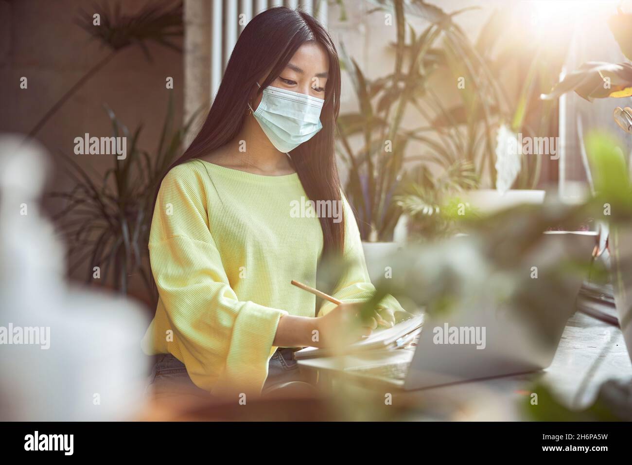 Attractive woman wearing yellow jumper while working in medical mask so as not to get virus at urban coffee shop Stock Photo