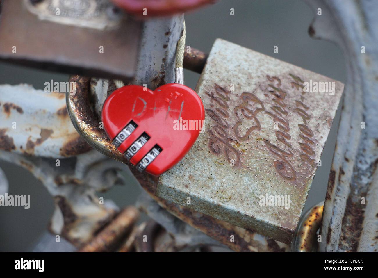 Red heart-shaped lock on the fence on the background of other castles, water, concrete pavement Stock Photo
