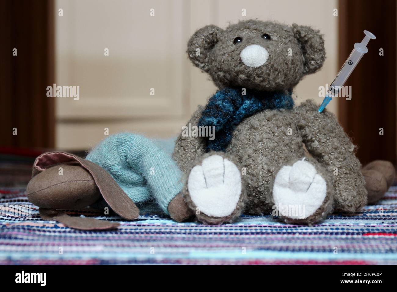 Zwei Kuscheltiere, ein liegender Hase und ein sitzender Teddy mit Spritze im Arm auf einem bunten Teppich. Stock Photo