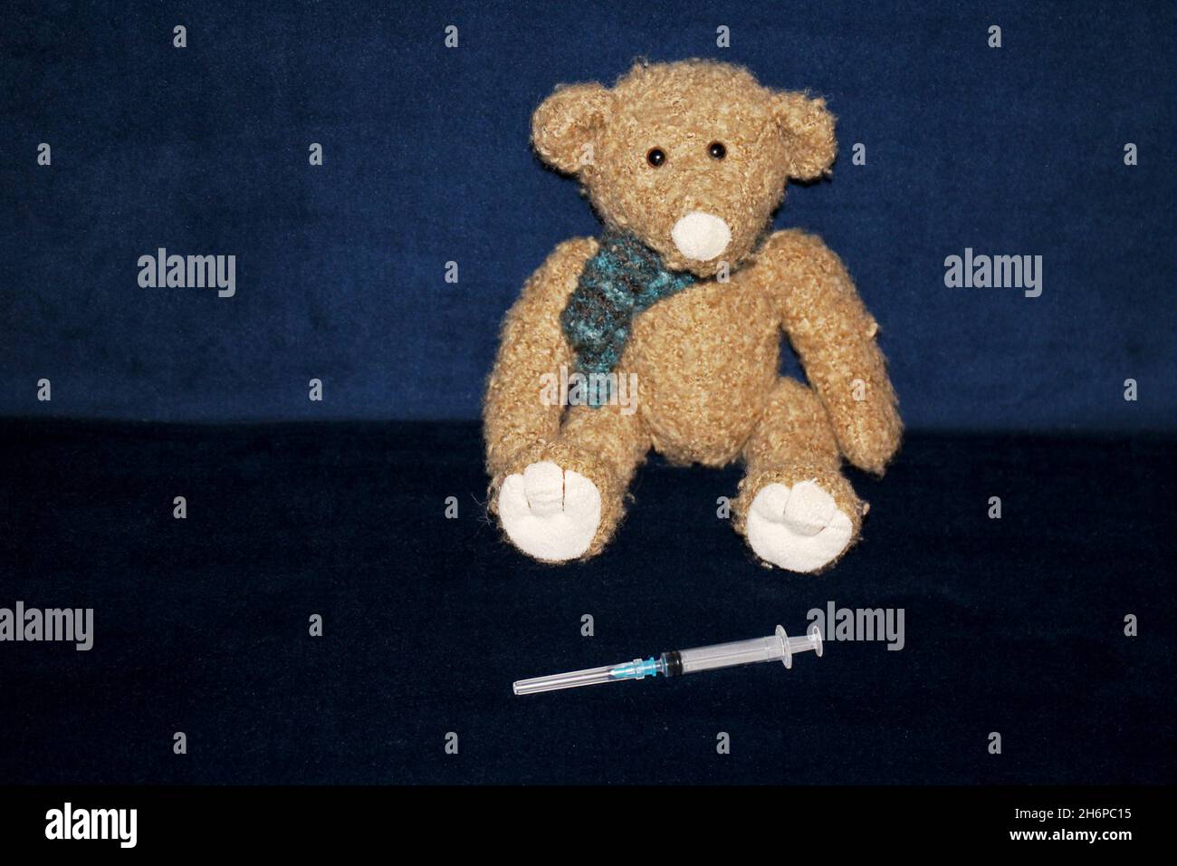 Kuscheltier Teddy sitzend auf einem dunkelblauen Sofa aus Samt. Vor ihm liegt eine Spritze. Stock Photo
