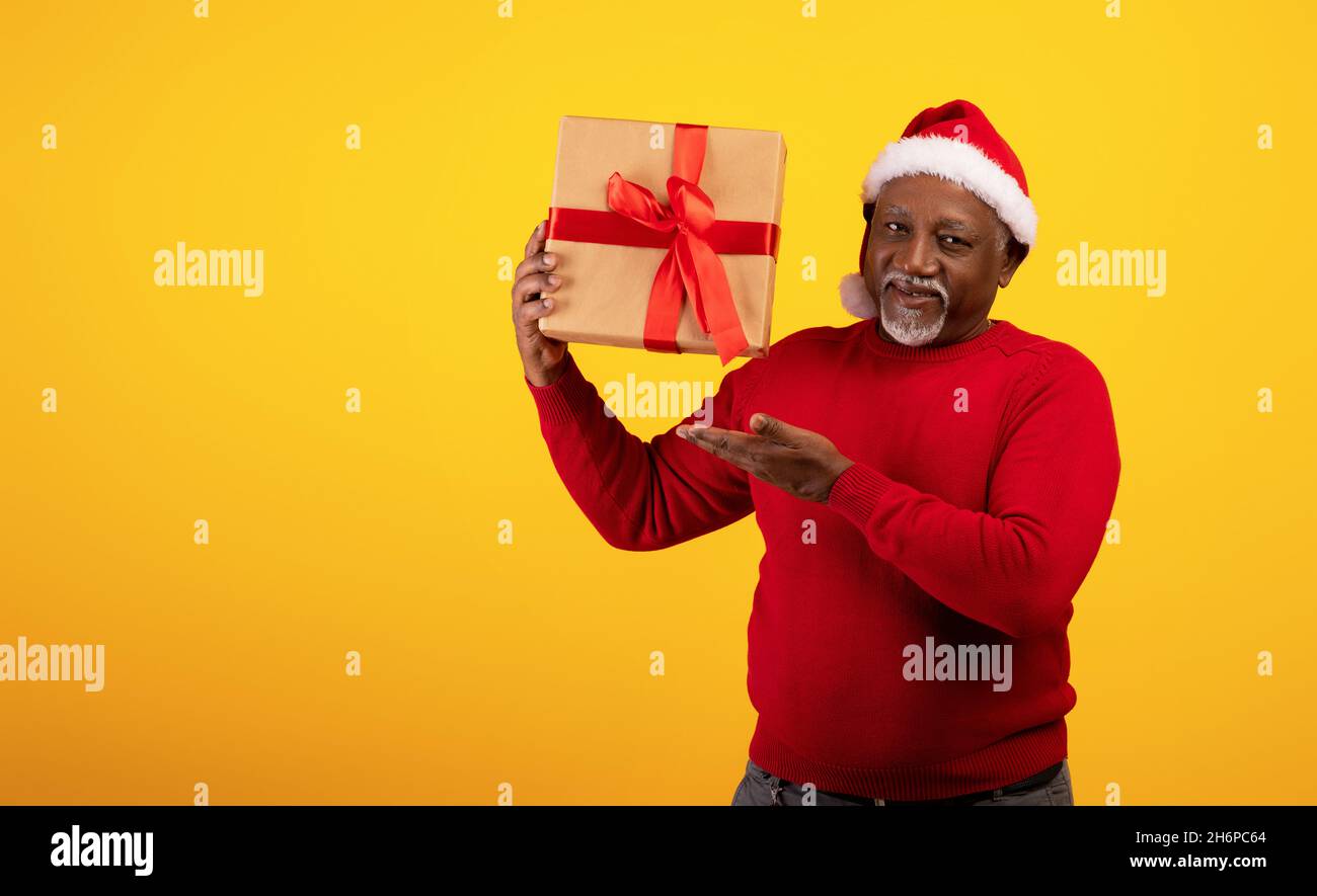 Christmas portrait of joyful senior black man in Santa hat holding Xmas gift over orange background, empty space Stock Photo