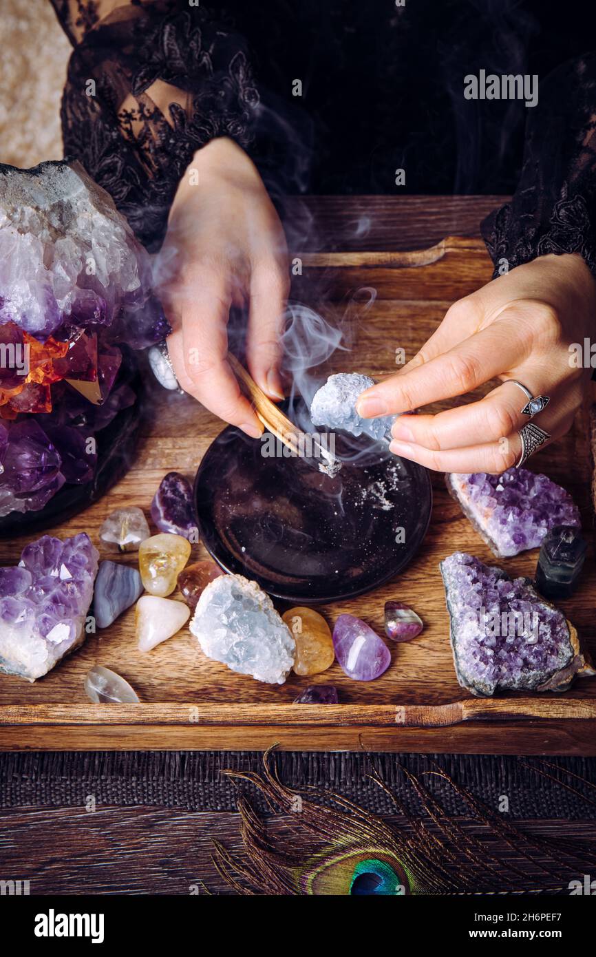 Glamorous woman in black dress cleansing crystals gemstones by smudging Palo Santo wood stick. Remove negative energy. Stock Photo