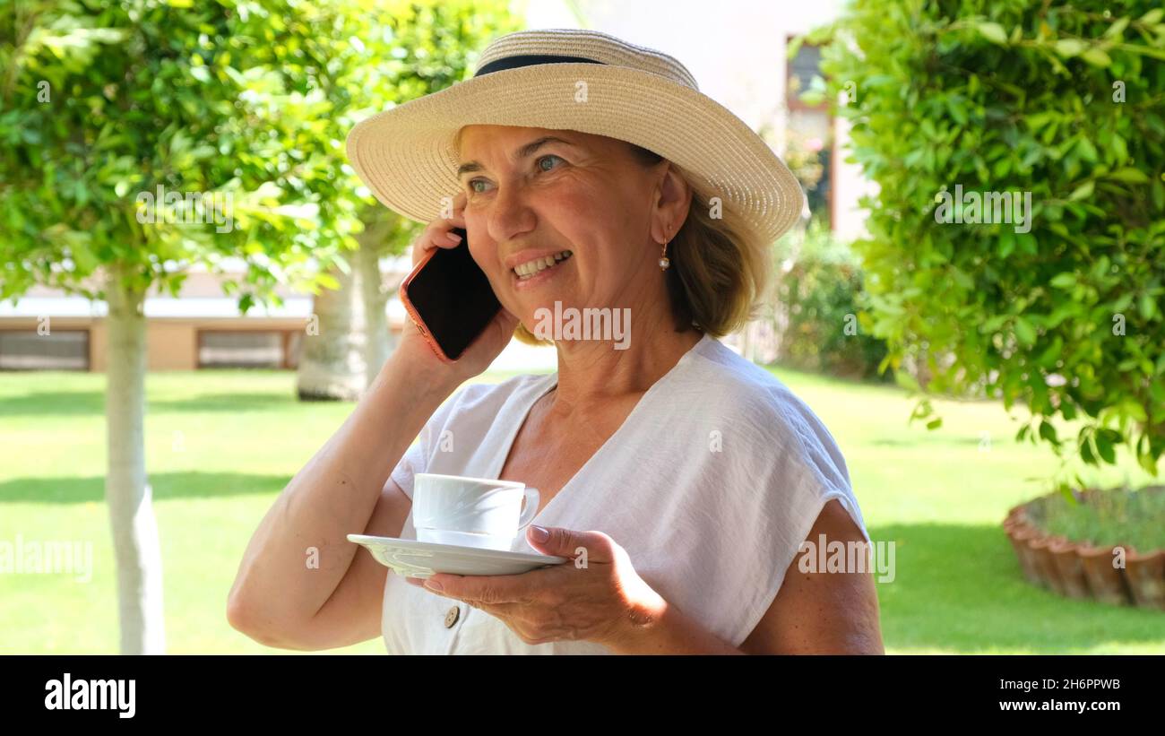 blonde senora woman, 50-55 years old, in a hat and a white dress, talks using a smartphone and holds a cup of coffee in her hands on her terrace on a Stock Photo