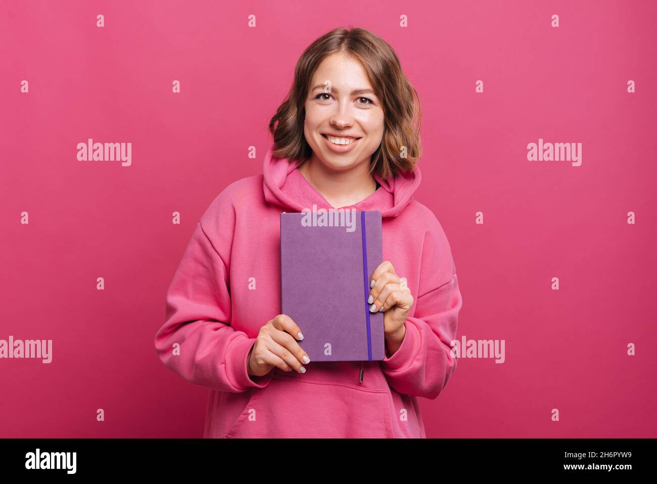 Portrait of young cheerful woman holding agenda planner over pink background. Stock Photo