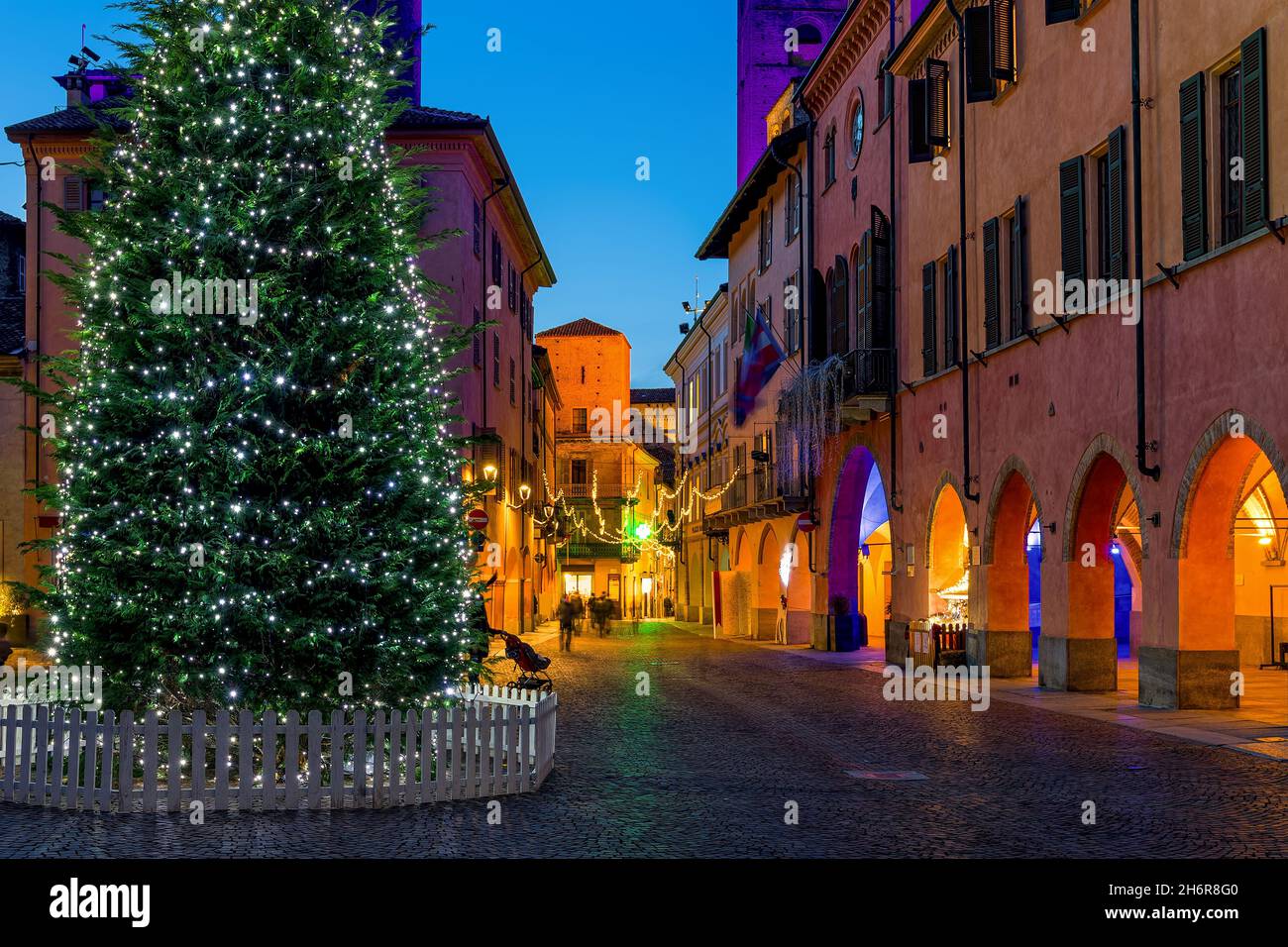 Illuminated Christmas tree on town square and cobblestone street among historic buildings in the evening in Alba, Piedmont, Northern Italy. Stock Photo