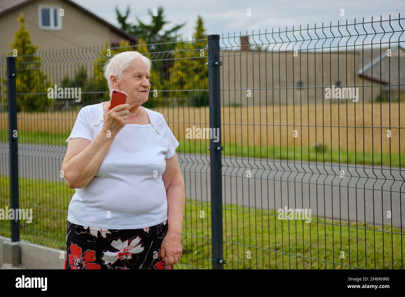 Half length view of an elderly woman talking on a cell phone while standing by the fence Stock Photo