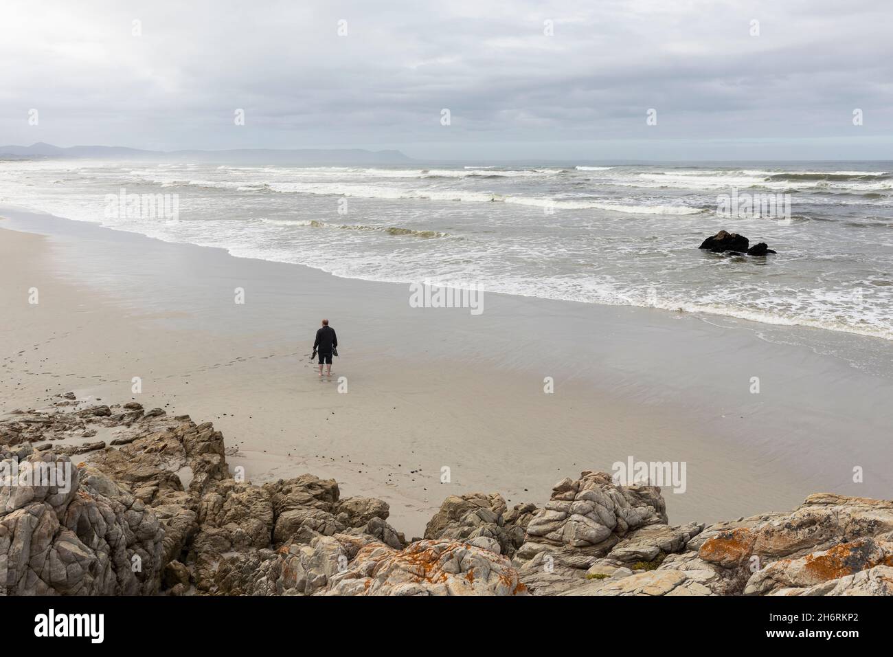 A man walking across sand to the water's edge on a beach, overcast day and surf waves breaking on shore. Stock Photo