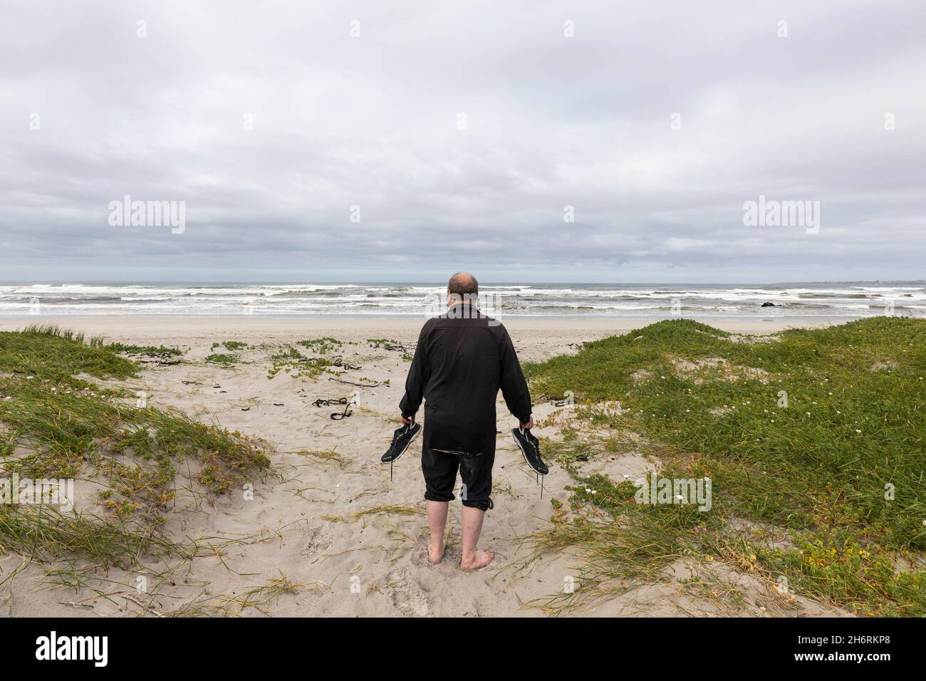 A mature man walking across a beach carrying his shoes in his hand Stock Photo