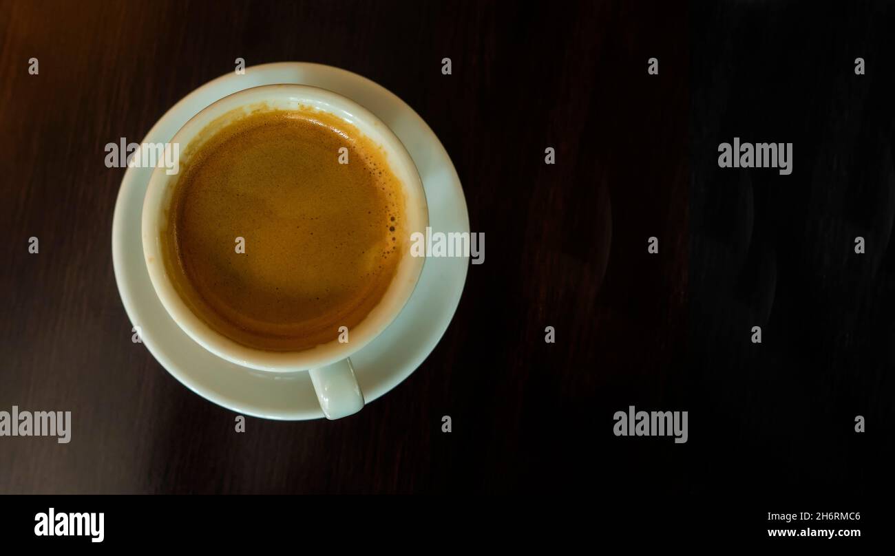 Black coffee with foam in white cup on saucer on dark background with