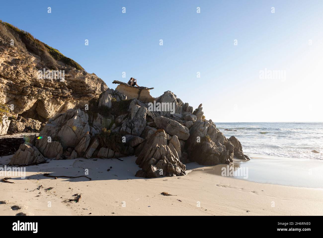 Two children perched on top of jagged rocks overlooking a sandy beach at low tide Stock Photo