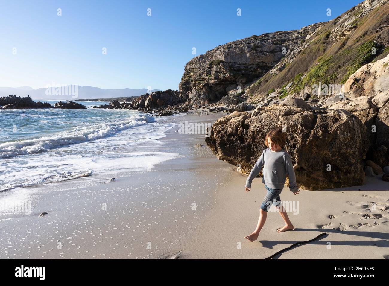 A young boy alone on a small stretch of sand under the cliffs by the ocean. Stock Photo