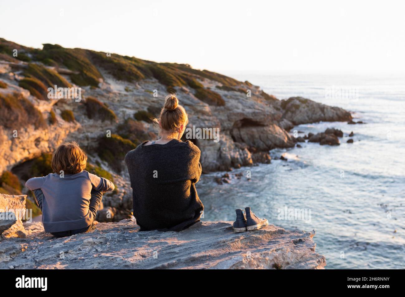 Teenage girl and young boy sitting on rocks looking over the sea at sunset Stock Photo