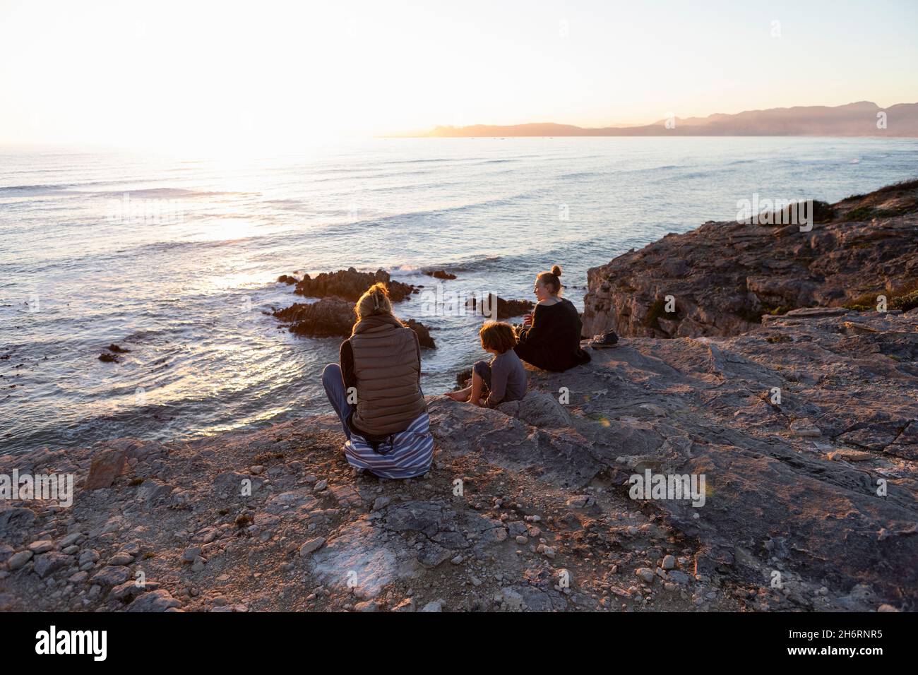 A family, mother and two children sitting watching the sunset over the ocean. Stock Photo