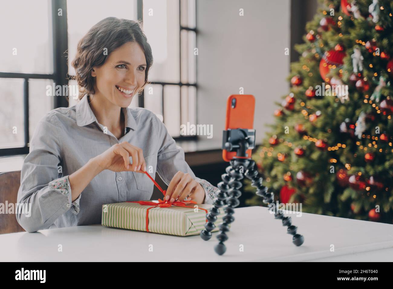 Joyful lady sitting at table with Xmas tree on background in front of phone on tripod Stock Photo
