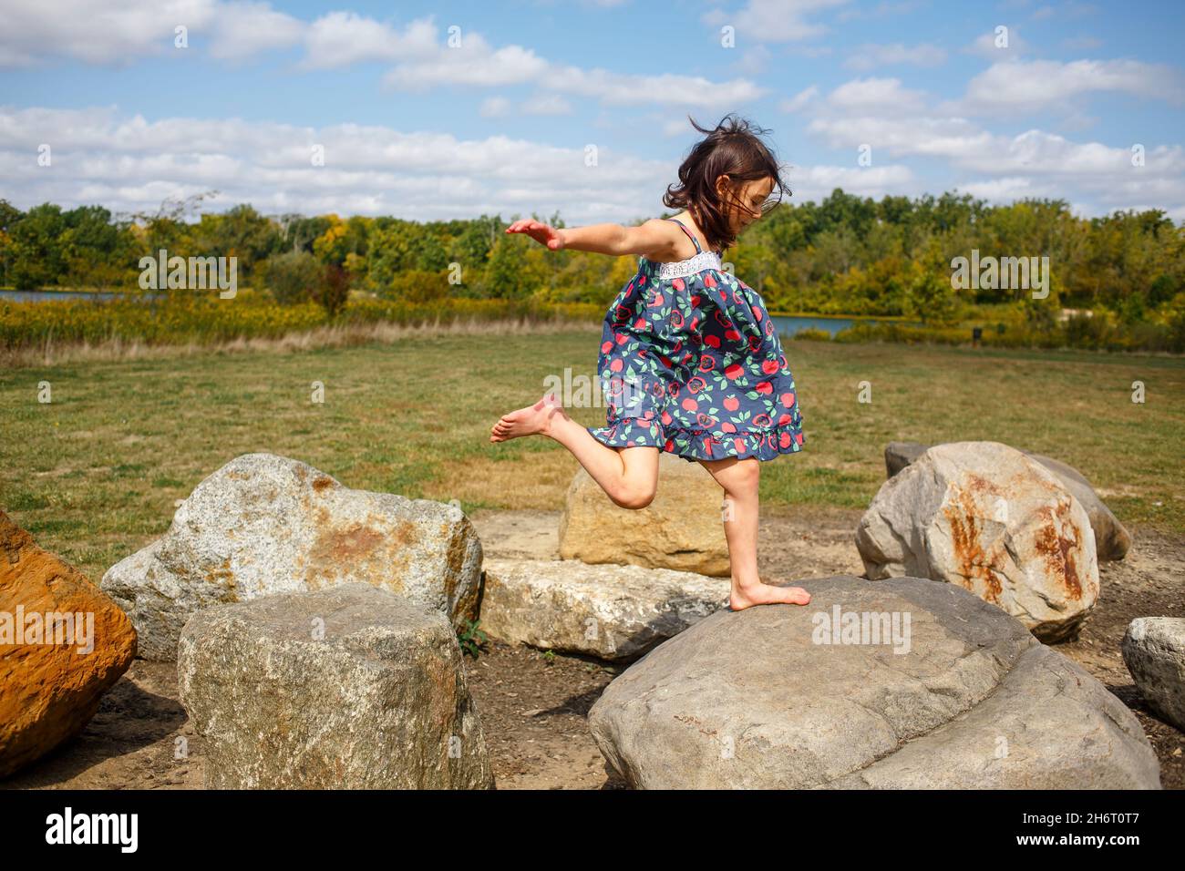 Small barefoot child in sundress leaps between large rocks Stock Photo