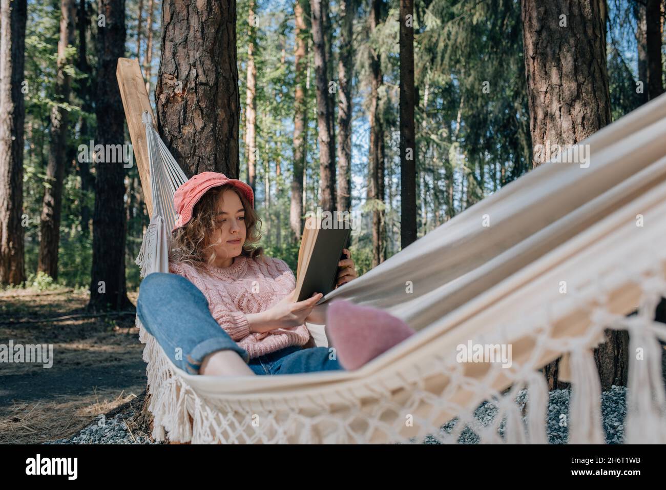 Young woman in hammock reading book in forest, relaxing. Stock Photo