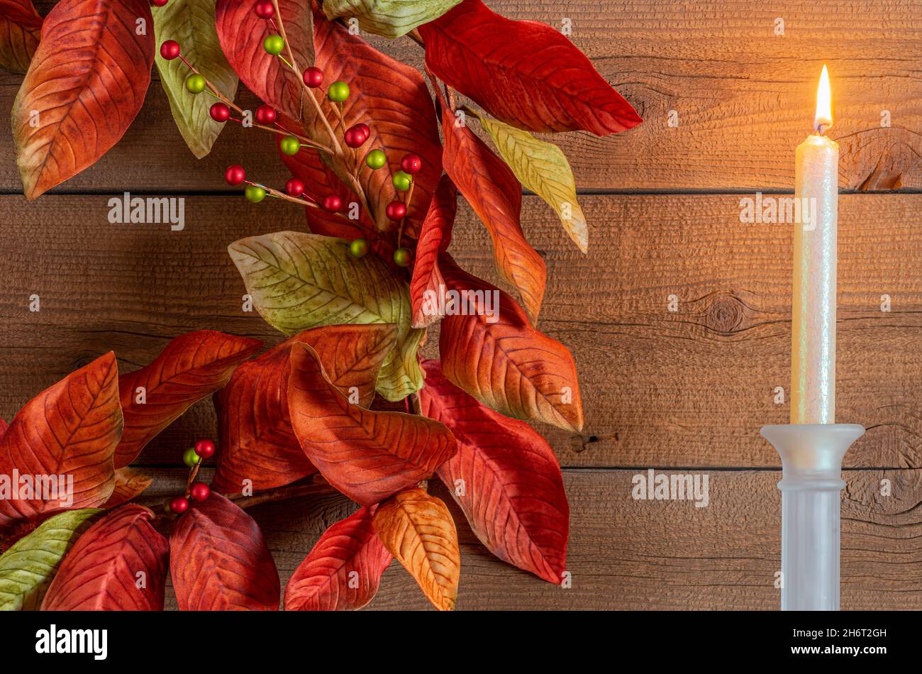 Colorful wreath in autumn and Christmas colors hangs next to a lit candle against heavily textured wood. Stock Photo