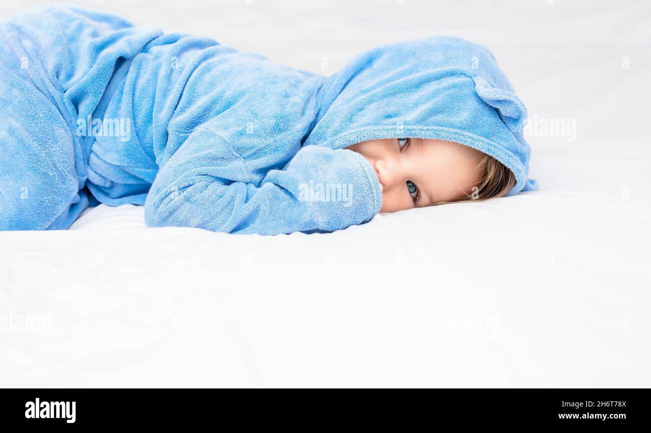 A child in a robe is lying on the bed. Baby in a blue towel on a white