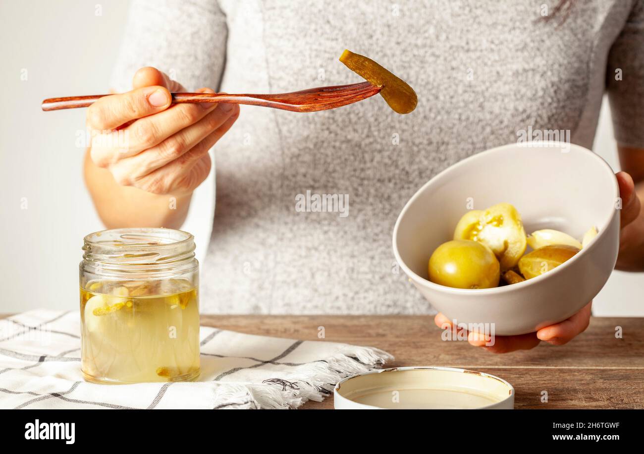 A young caucasian woman is eating homemade pickled vegetables. She is taking a cucumber from a bowl containing tomatoes and cucumbers. Stock Photo