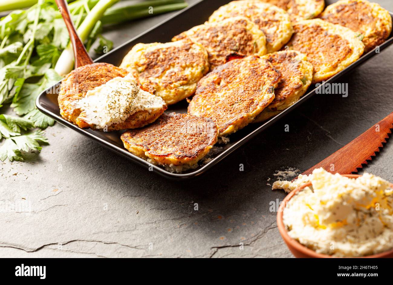 Turkish style zucchini pancakes or fritters known as mucver, served on black plate with fresh greens and sour cream. Close up image on dark stone back Stock Photo