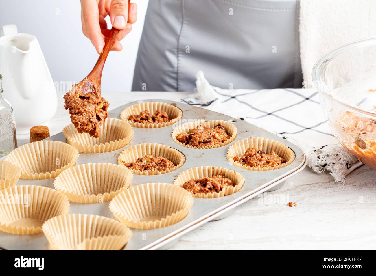 making of delicious fruit and nut cupcake on white marble countertop background. Muffin tin with liner, ingredients, and utensils are seen. A woman is Stock Photo