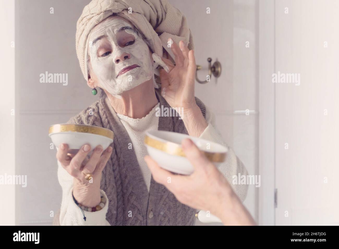 lderly woman with towel on her head smiles and applies cleansing mask . Stock Photo