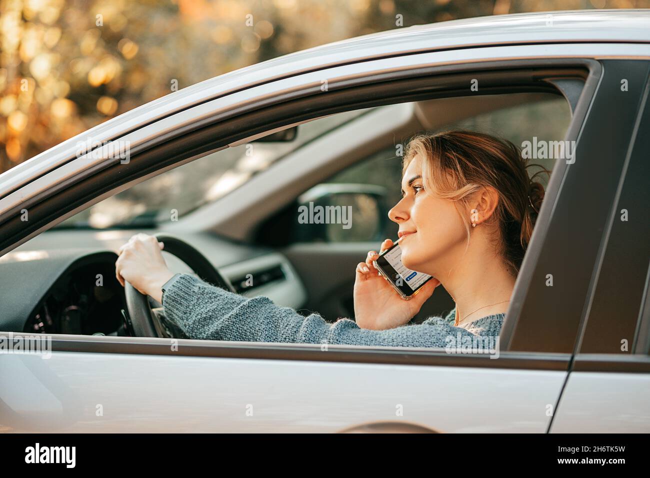Photo of Young beautiful blonde woman drives car, violates traffic rules, gets distracted, looks at screen of mobile phone, uses smartphone, fast pace Stock Photo