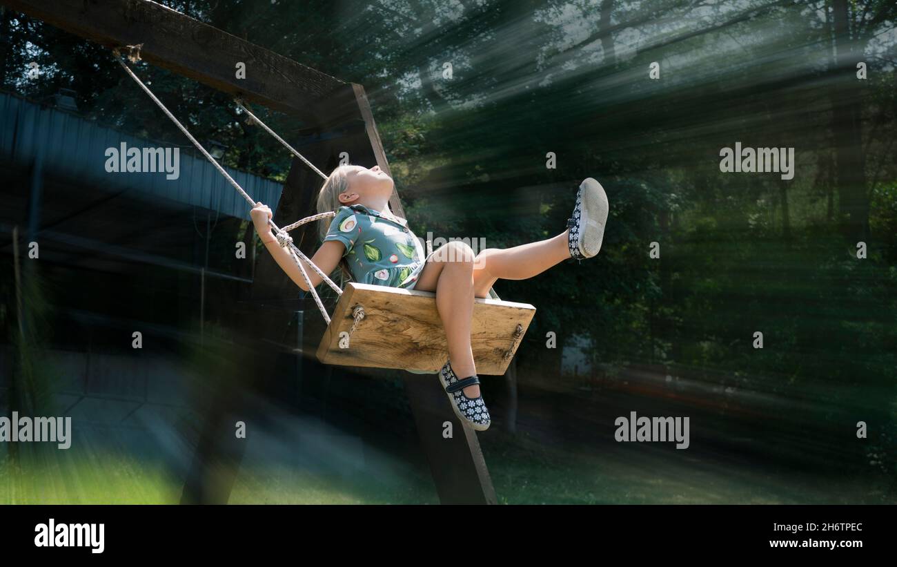 Beautiful little girl in flight on a swing against the background of a blurred in motion park with large trees in the sun's rays. Joyful, carefree, ha Stock Photo