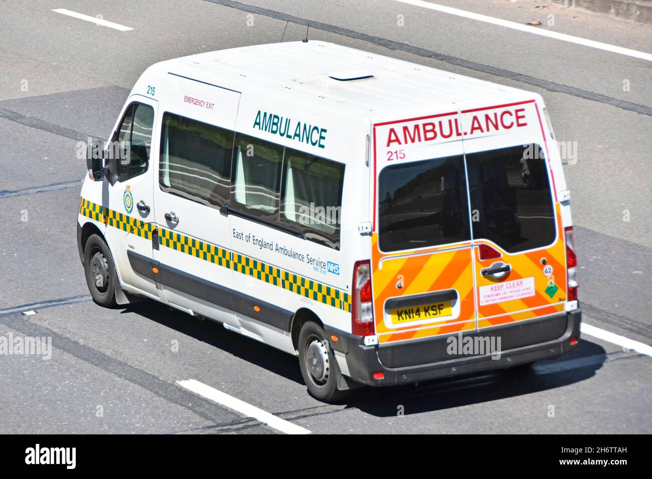 Side back & aerial view East of England patient transfer type of NHS healthcare ambulance with high visibility vehicle markings driving on uk motorway Stock Photo