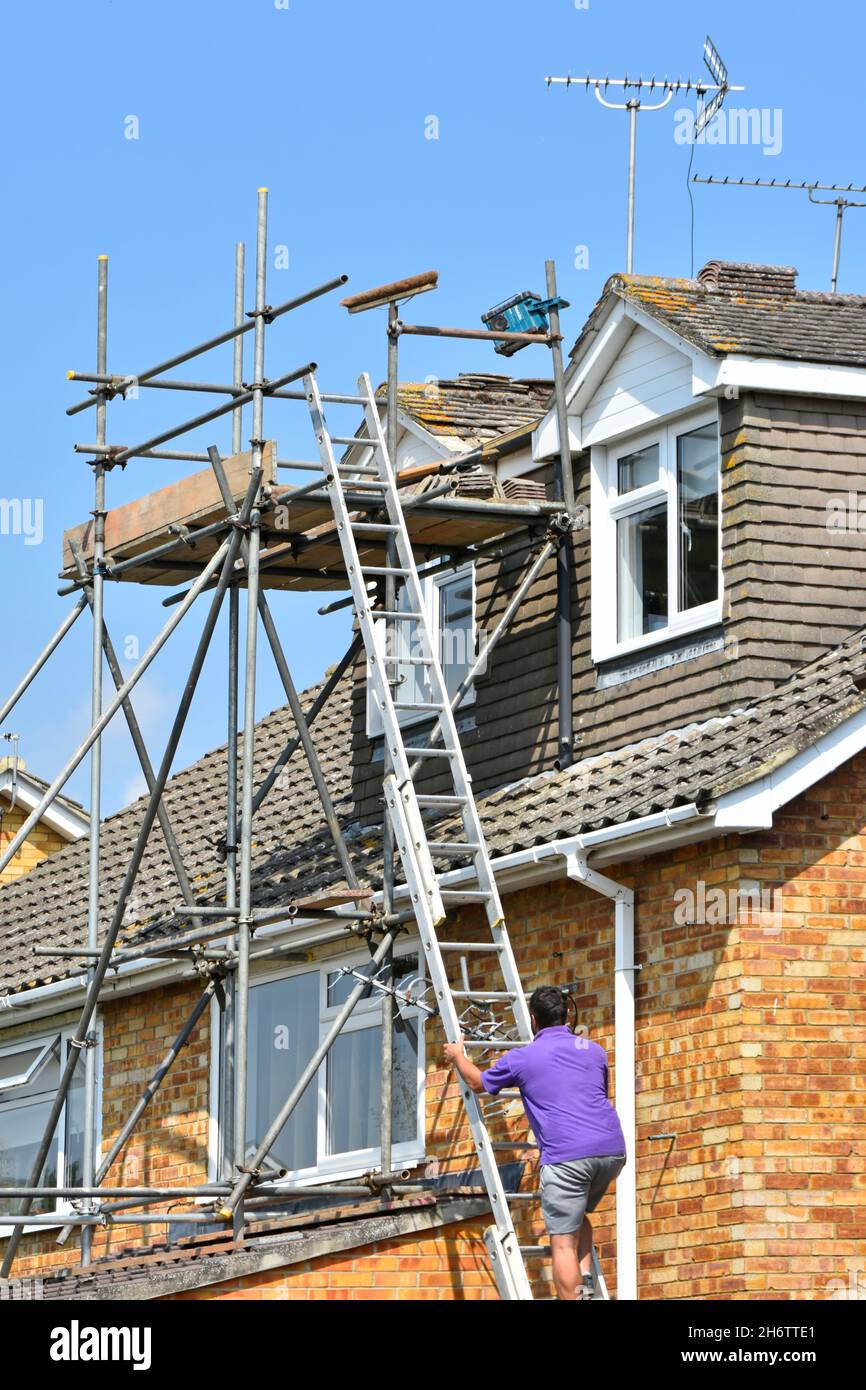 Builder man climbing up ladder on scaffold tower around dormer rooms for roof maintenance repair & refit plain roof ridge tiles to house England UK Stock Photo