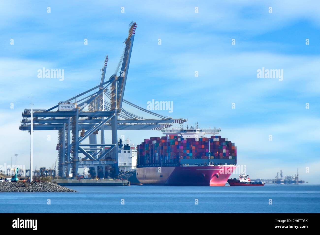 Tugboat pushing big container ship & full load of shipping containers to birth below giant tall cranes at London Gateway Thames estuary port Essex UK Stock Photo