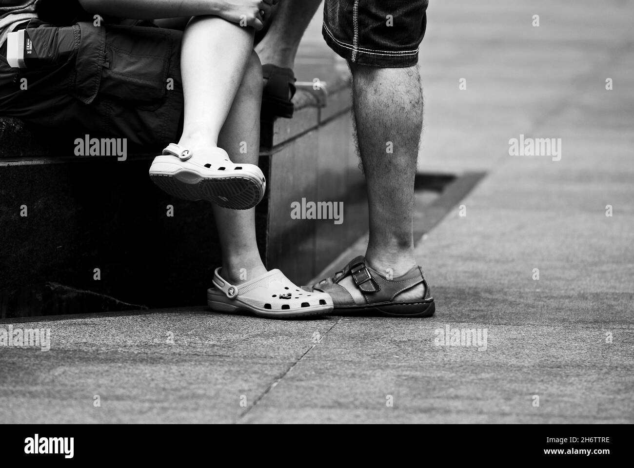 Greyscale shot of the legs of two males sitting on a bench outdoors Stock Photo