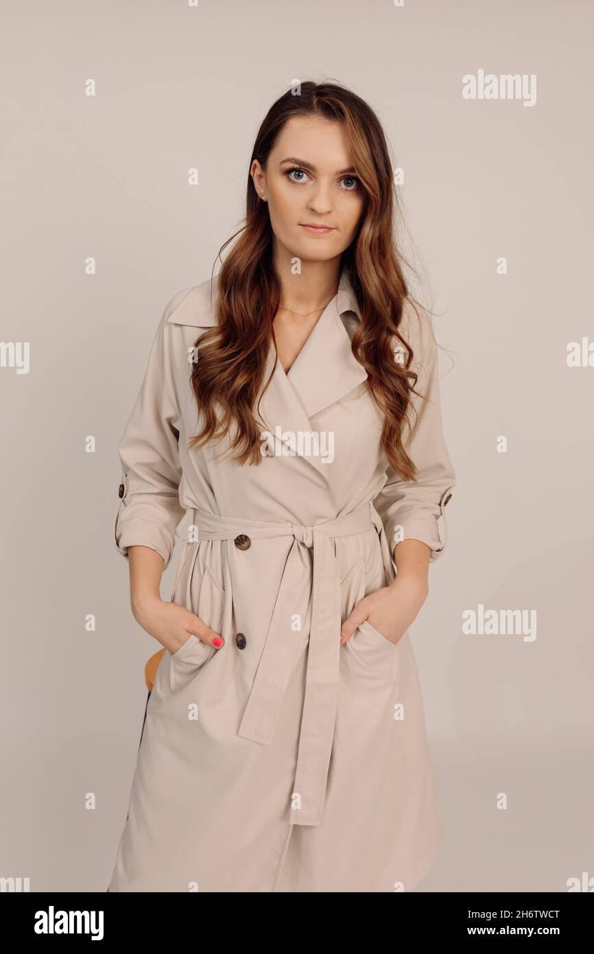 Young beautiful cute serios long brown wavy hair woman top manager in beige trench coat looks at camera, smiles positively, keeps her hands in pockets Stock Photo