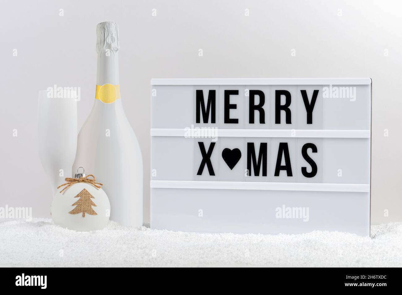 White champagne bottle, champagne glass and lightbox with text MERRY XMAS on a table with snow. Minimal Christmas or New Year holiday concept. Stock Photo