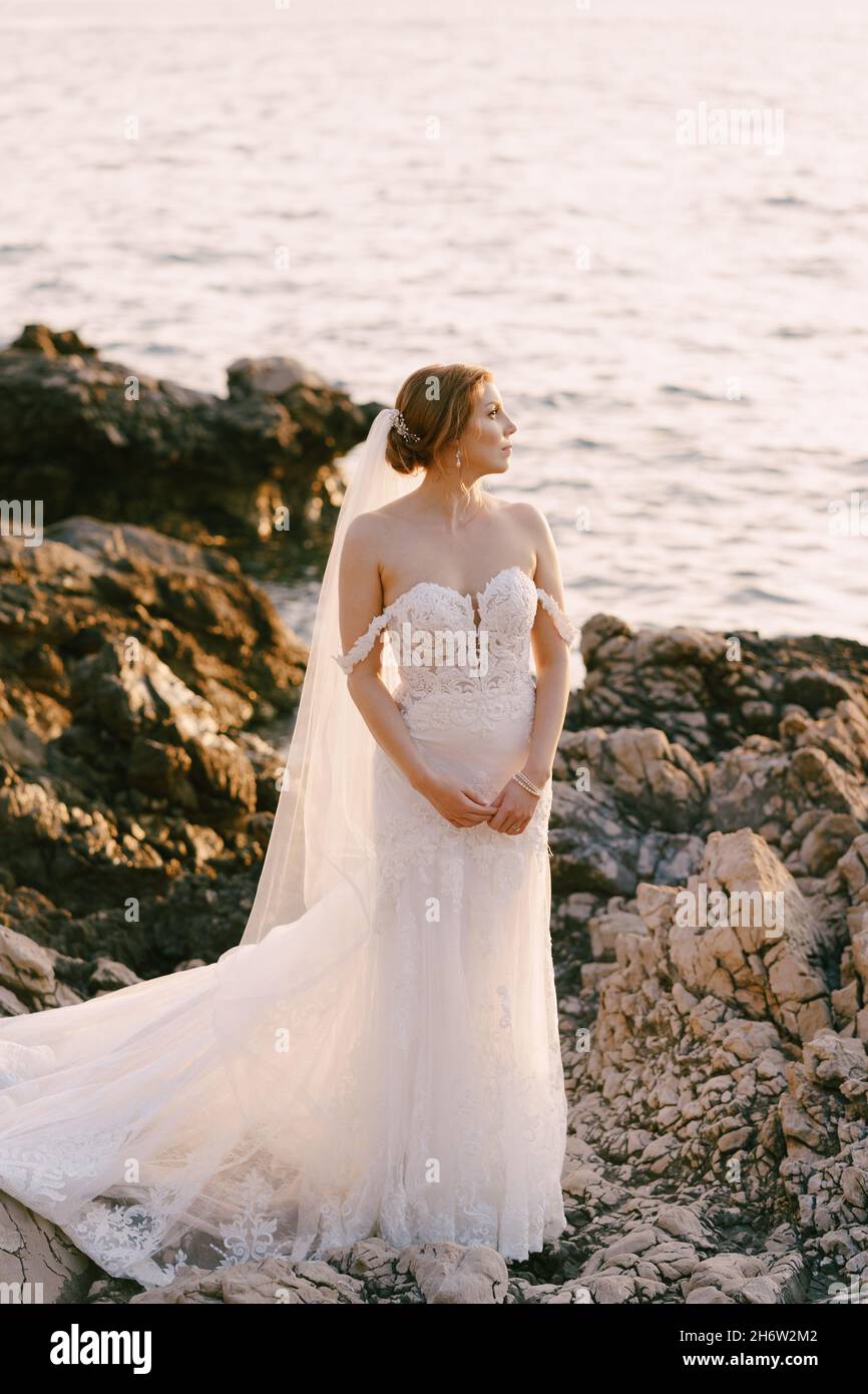 Bride in a white dress and veils stands on the rocks with her back to ...