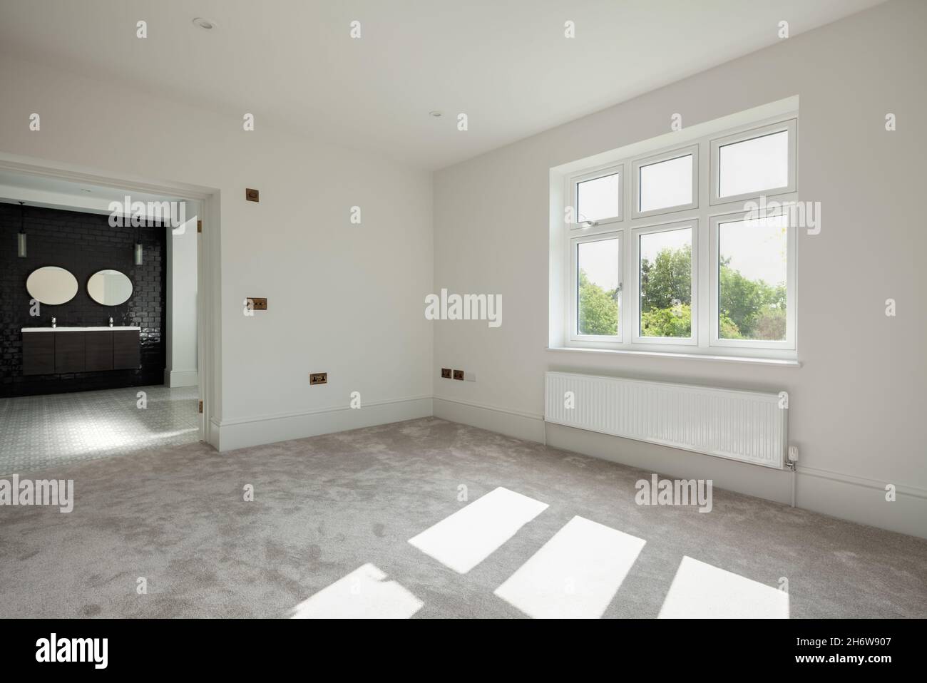 Wratting, England - August 19 2019: Empty sunlit bedroom space with doors opening onto ensuite tiled bathroom simply decorated in white and carpeted Stock Photo
