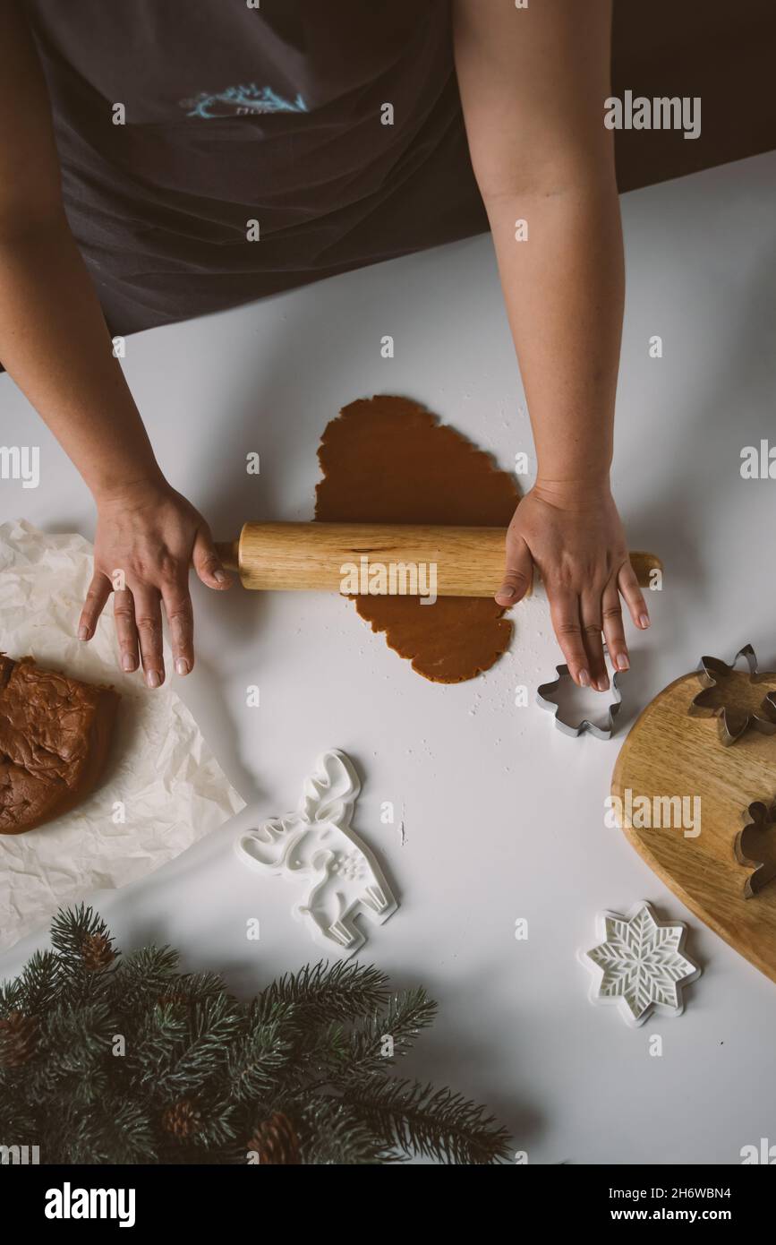 Female hands roll out raw ginger dough with a rolling pin on a white table. The process of preparing Christmas gingerbread cookies. Top view, headless Stock Photo