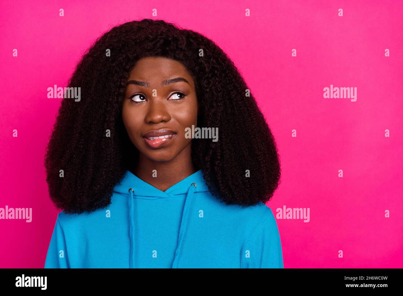 Photo of funny doubtful dark skin woman dressed blue sweatshirt biting lip looking empty space isolated pink color background Stock Photo