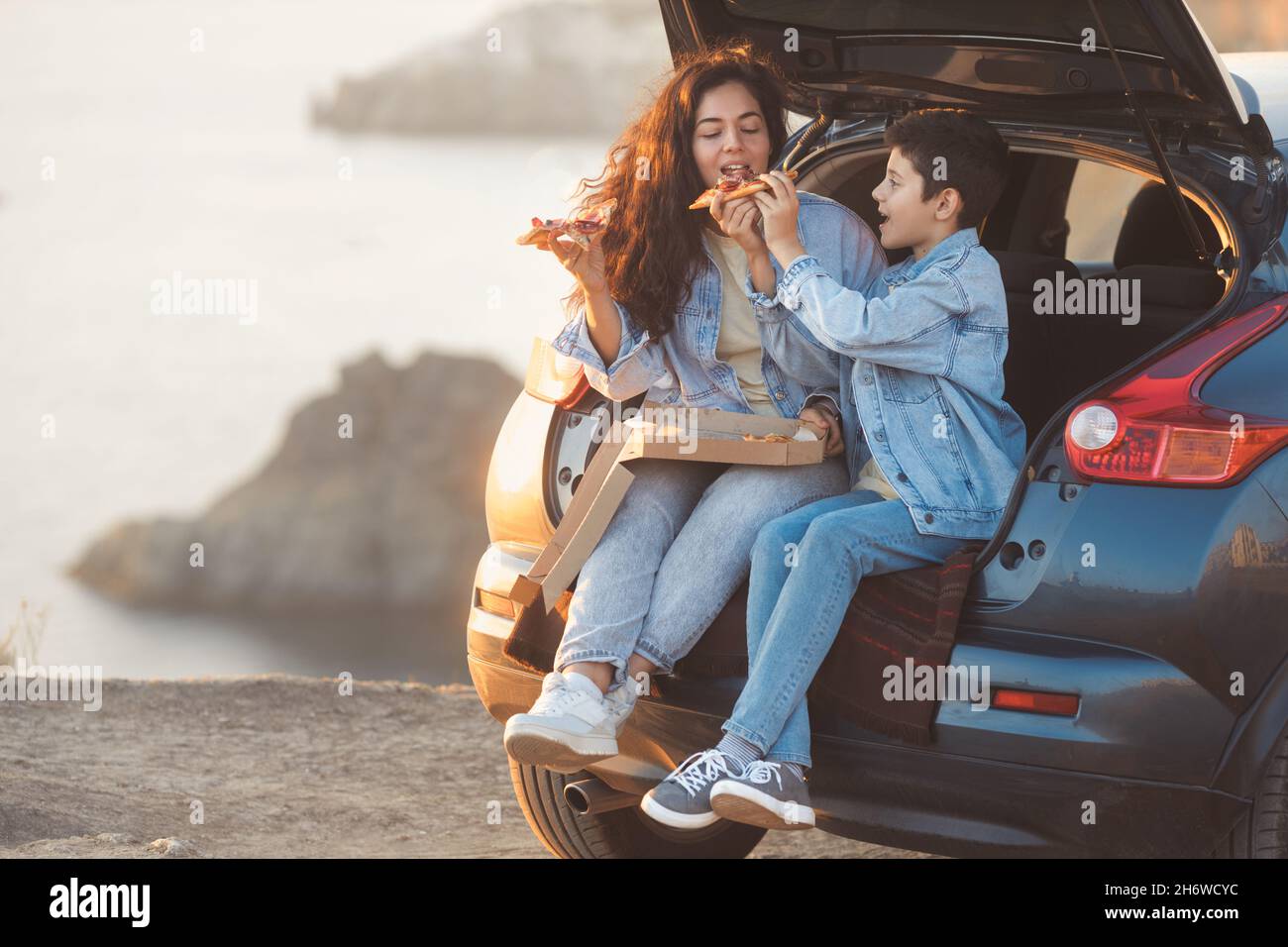 A young mother and her teenage son in denim clothes sitting in the open trunk of a car and eating pizza Stock Photo