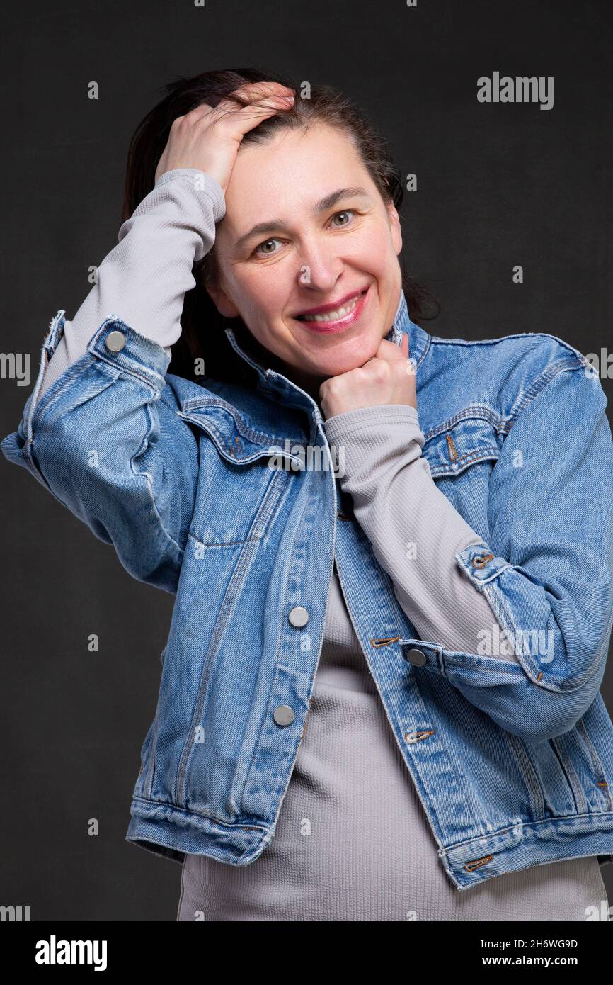 Happy fifty year old woman with dark hair in a denim jacket on a gray
